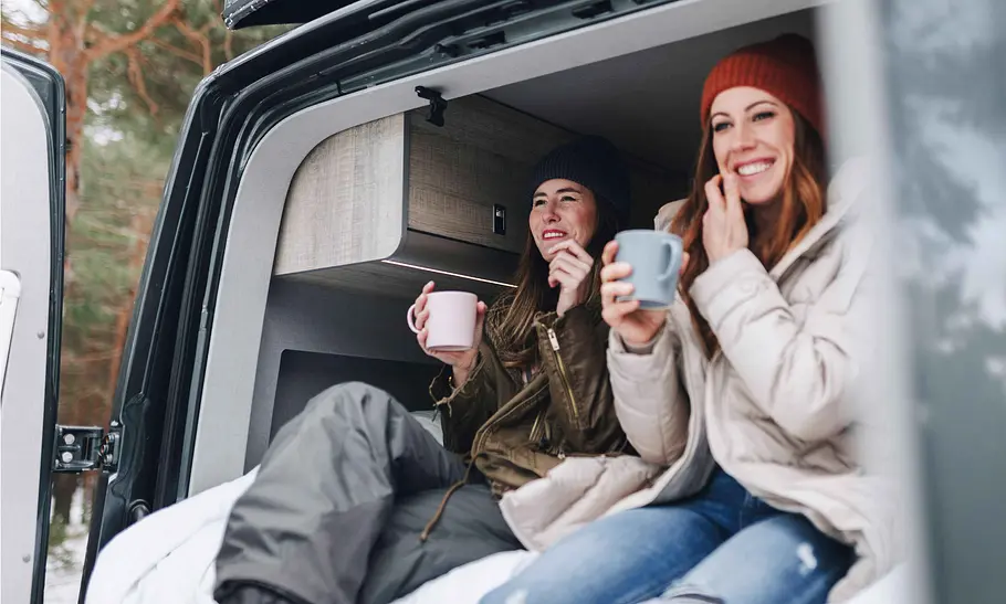 Zwei junge Frauen im Outdoor-Outfit sitzen im Heck eines Campingbusses und trinken Kaffee.