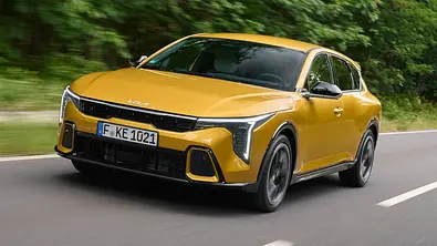 Der Kia K4 fahrend von schräg vorne links auf einer Landstraße in einem Waldstück. - Foto: Kia