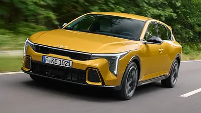 Der Kia K4 fahrend von schräg vorne links auf einer Landstraße in einem Waldstück. - Foto: Kia
