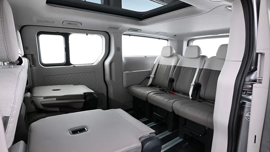 Kia PV7 Passenger Blick durch die geöffnete linke Schiebetüre auf die dritte Sitzreihe im Fahrgastraum des Kia PV7 Passenger mit umgelegten Einzelsitzen in der zweiten Reihe.