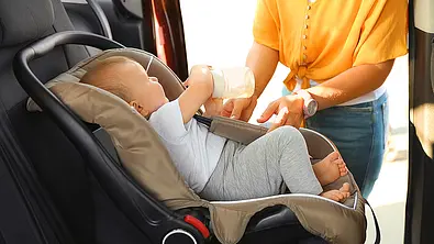 Das Bild zeigt ein Kleinkind in einem vorwärtsgerichteten Reboarder auf der Rückbank eines Autos. Das Kind hält eine Babyflasche in der Hand, eine Person steht an der geöffneten Schiebetüre des Autos und schnallt das Kind an. - Foto: Imago
