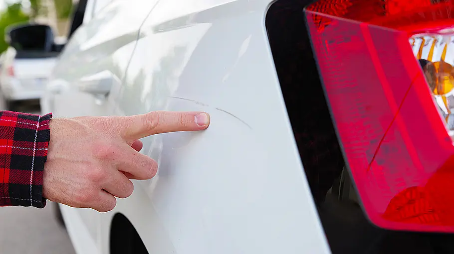 Kratzer am Auto entfernen Eine Person zeigt mit dem Zeigefinger auf einen Kratzer im Lack über dem hinteren linken Radlauf eines weißen Skoda Fabia.