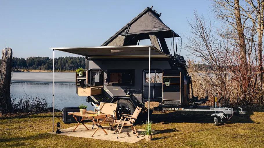 Die Außenaufnahme zeigt den abgestellten Kuckoo Camper Max mit ausgezogenem Aufstelldach und Markise an einem See.