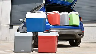 Die sechs getesteten Auto-Kühlboxen von vorne nebeneinander stehend fotografiert. Dahinter drei Kühlboxen in einem geöffneten Kofferraum. - Foto: Alexander Koch Die sechs getesteten Auto-Kühlboxen von vorne nebeneinander stehend fotografiert. Dahinter drei Kühlboxen in einem geöffneten Kofferraum. - Foto: Alexander Koch