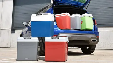Die sechs getesteten Auto-Kühlboxen von vorne nebeneinander stehend fotografiert. Dahinter drei Kühlboxen in einem geöffneten Kofferraum. - Foto: Alexander Koch