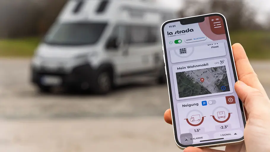 Das Bild zeigt ein Smartphone mit der La Strada-App in der Hand eines Testers. Im Hintergrund parkt ein La Strada-Wohnmobil.