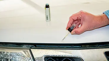 Eine Person bessert einen Steinschlag auf der Motorhaube eines weißen Autos mit einem Lackstift aus. - Foto: Imago Eine Person bessert einen Steinschlag auf der Motorhaube eines weißen Autos mit einem Lackstift aus. - Foto: Imago