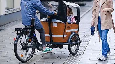 Das Bild zeigt ein stehendes Lastenrad mit Kindern im Ladeabteil vor dem Lenker. Eine Person sitzt am Lenker des Fahrrads, eine zweite Person läuft am Fahrrad vorbei. - Foto: Imago