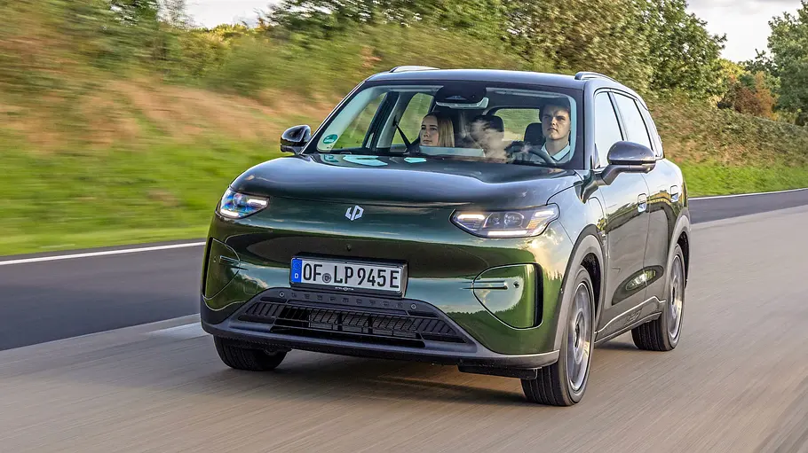 Ein dunkelgrüner Leapmotor B03X mit zwei sichtbaren Insassen fährt auf einer Straße, im Hintergrund stehen Bäume und Sträucher.