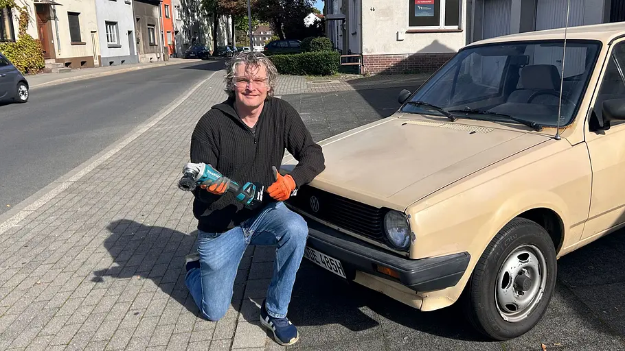 Makita Akku-Flex DGA504 Ein Produkttest-Redakteur kniet mit Makita-Winkelschleifer vor dem Heck eines Oldtimers (VW Polo 86 C), der als Versuchsobjekt dient.