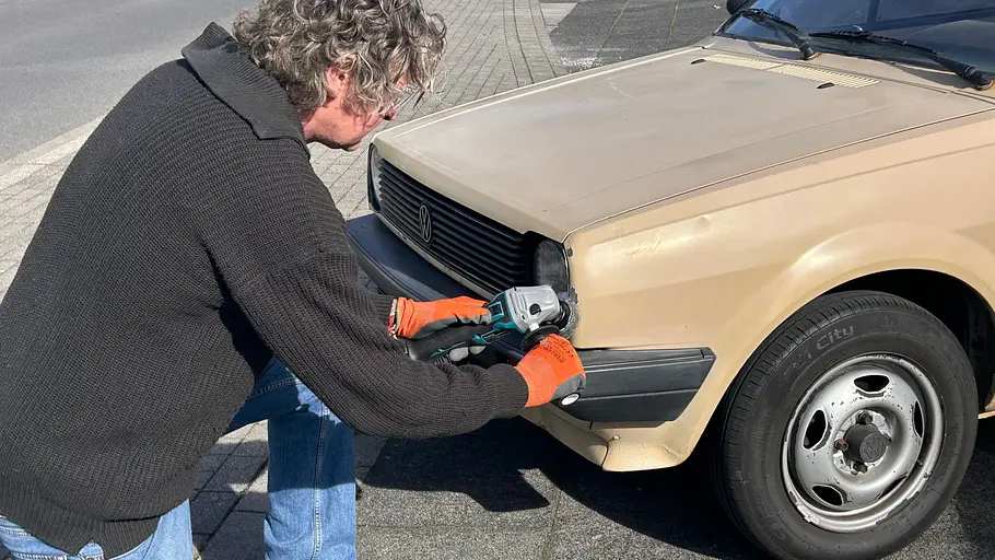 Makita Akku-Flex DGA504 Ein Produkttest-Redakteur kniet mit Makita-Winkelschleifer vor dem Heck eines Oldtimers (VW Polo 86 C), der als Versuchsobjekt dient.