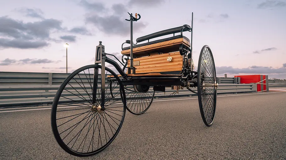 Ein Nachbau des Fahrzeugs mit Gasmotorenbetrieb von Carl Benz von 1896 steht vor der Leitplanke einer modernen Rennstrecke.