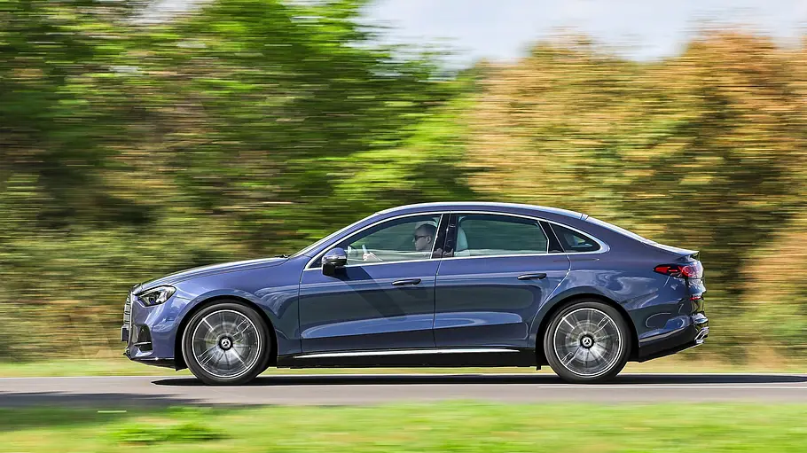 Test Mercedes C 400 4Matic elektrisch Eine blaue Mercedes C-Klasse elektrisch fährt auf einer Landstraße an grünen Bäumen und bewaldeten Hügeln vorbei.