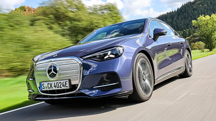 Test Mercedes C 400 4Matic elektrisch Eine blaue Mercedes C-Klasse elektrisch fährt auf einer Landstraße an grünen Bäumen und bewaldeten Hügeln vorbei.