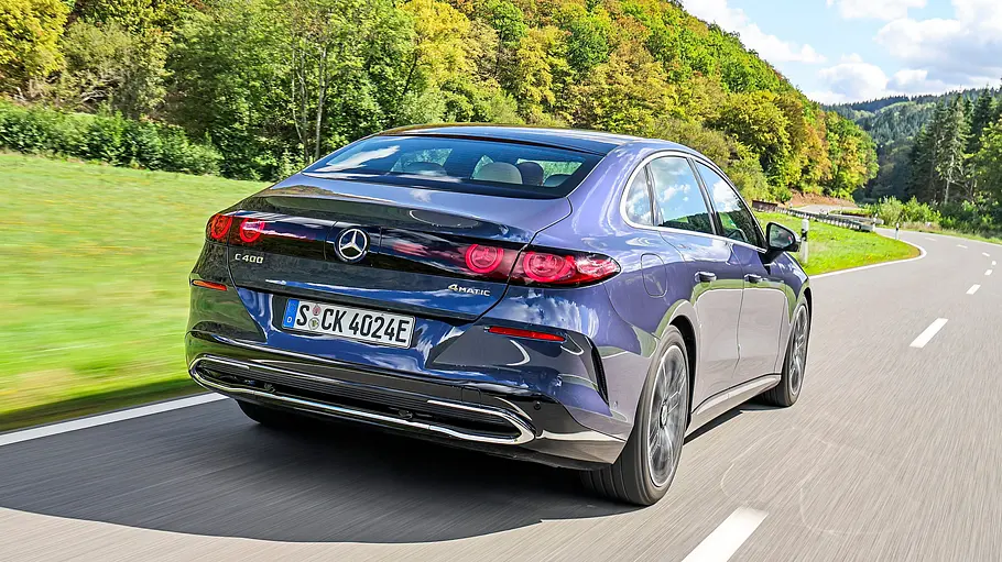 Test Mercedes C 400 4Matic elektrisch Eine blaue Mercedes C-Klasse elektrisch fährt auf einer Landstraße an grünen Bäumen und bewaldeten Hügeln vorbei.