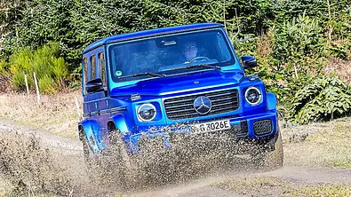 Blick von schräg vorne auf den fahrenden Mercedes G 580 EQ, der gerade durch eine tiefe Matschpfütze fährt. - Foto: Zibigniew Mazar