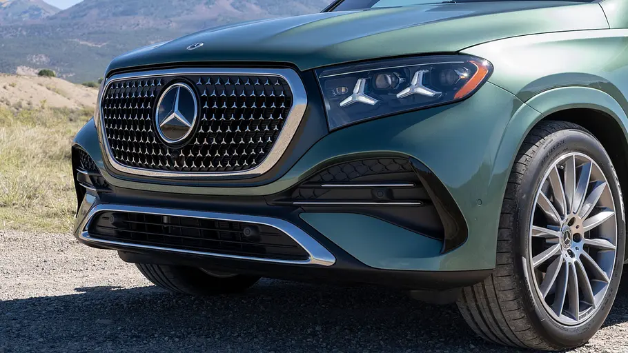 Mercedes GLE 580 4Matic Modellpflege Fahrbericht Ein grüner Mercedes GLE steht auf Schotter vor Bergen. Die Nahaufnahme zeigt Kühlergrill, Scheinwerfer und Vorderrad.