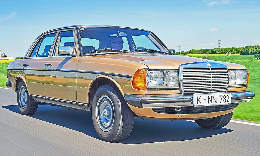 Ein fahrender Mercedes W123 von vorne. Im Hintergrund sieht man eine grüne Fläche.