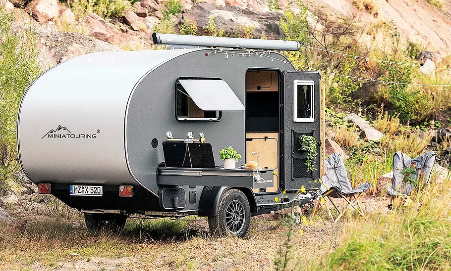 Das Bild zeigt den Miniatouring M24 (2025) stehend von schräg hinten rechts fotografiert. Die Türe ist geöffnet, der Küchenauszug ist ausgefahren und neben dem Wohnwagen stehen Campingmöbel.