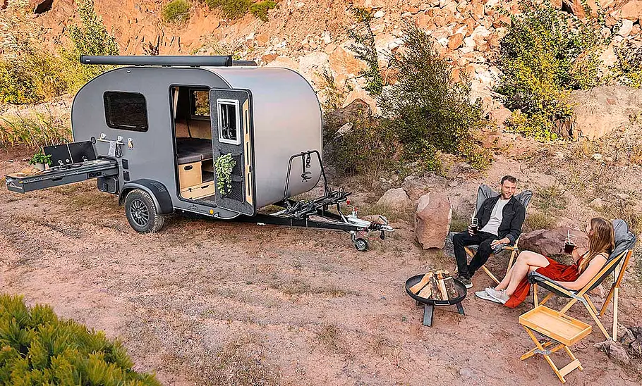 Das Bild zeigt den Miniatouring M24 (2025) stehend von schräg vorne rechts fotografiert. Die Türe ist geöffnet und der Küchenauszug ist ausgefahren. Auf Campingmöbeln sitzen zwei Personen um eine Feuerschale.