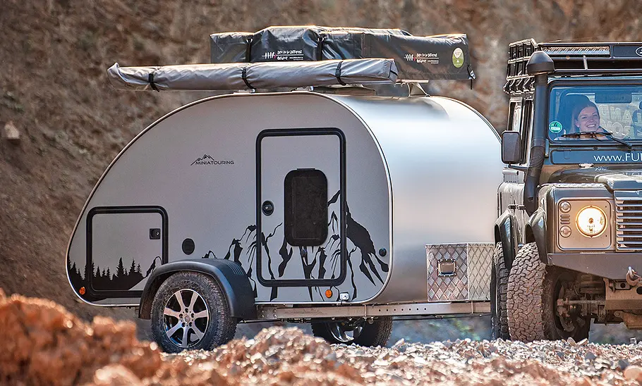 Miniatouring Mini-Caravan M22 (2026) hängt an einem Land Rover Defender und fährt durchs Gelände. Ansicht des Caravan von rechts.