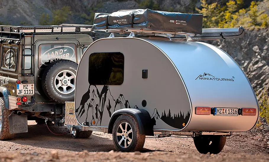 Miniatouring Mini-Caravan M22 (2026) hängt an einem Land Rover Defender und fährt durchs Gelände. Ansicht des Caravan von links.