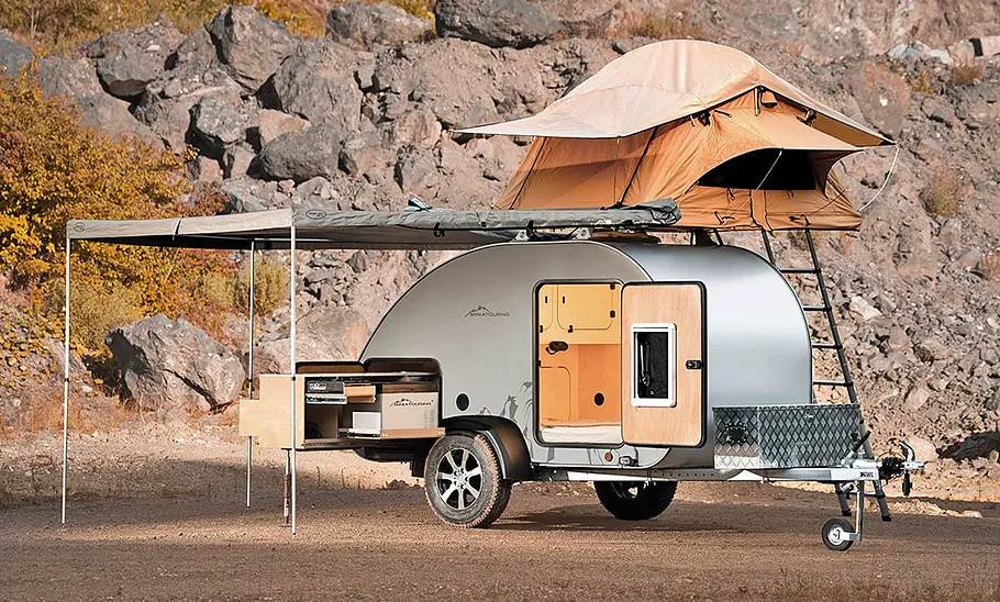 Der Miniatouring Mini-Caravan M22 (2026) steht im Gelände, ohne Zugfahrzeug, Ansicht von rechts. Die Tür des Wohnwagens ist geöffnet, die Küchenzeile herausgezogen, die Markise ist aufgebaut und es ist ein Dachzelt auf dem Dach ausgeklappt.