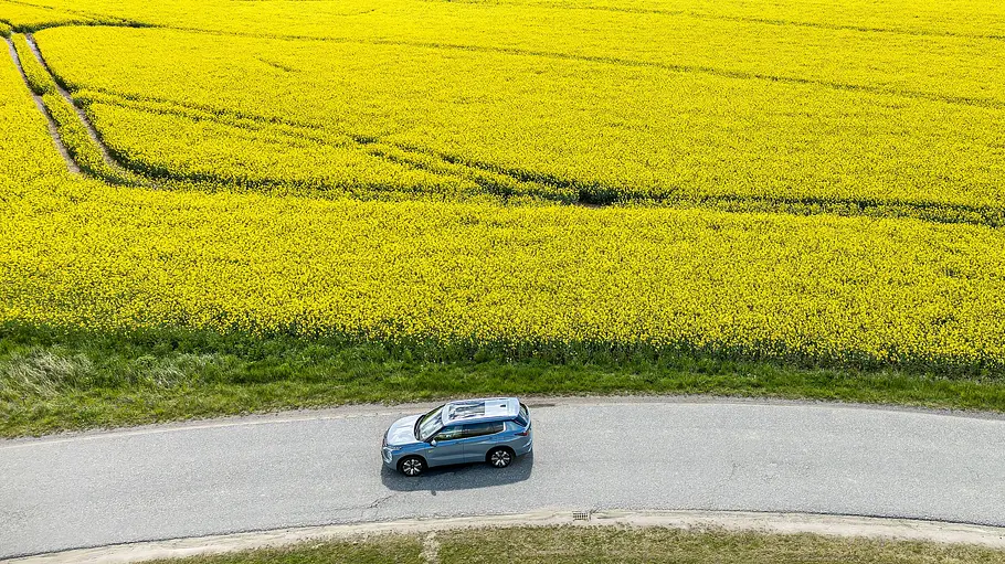 Drohnen-Aufnahme: Mitsubishi Outlander fährt auf einer Landstraße, im Hintergrund gelb blühende Felder.