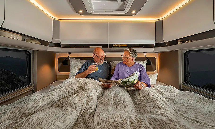 Das Bild zeigt das Heckbett des Morelo Loft (2024). Zwei Personen sitzen bettfertig auf dem Bett und lesen in einem Magazin.
