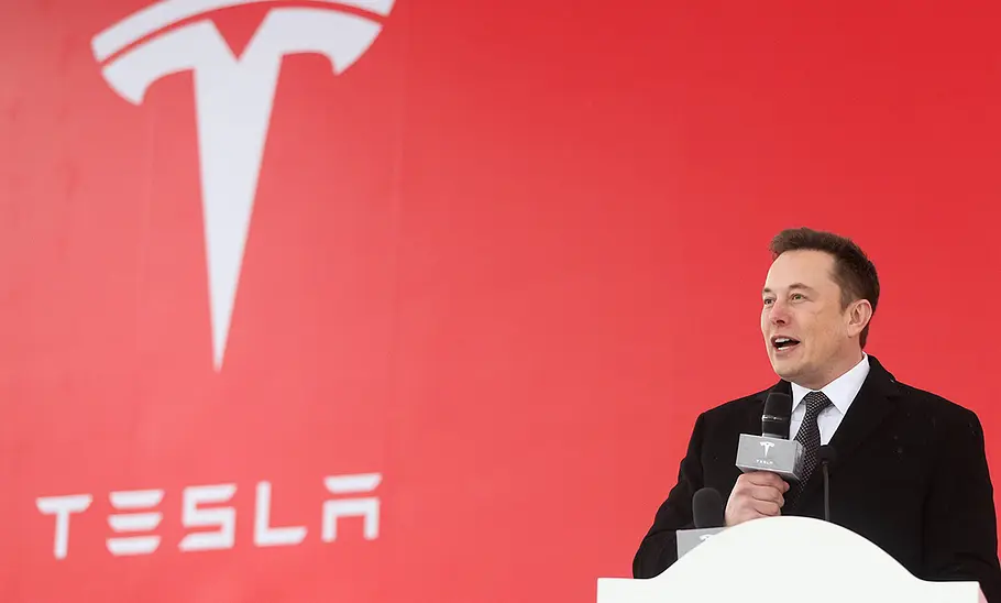 Elon Musk am Rednerpult, dahinter das Tesla-Logo auf einer großen roten Wand.