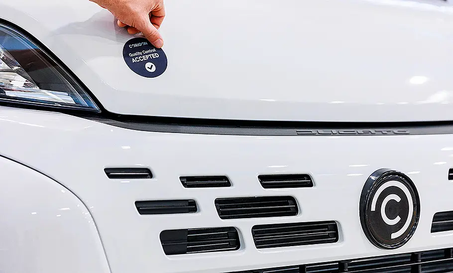 Neue Hymer-Marke Corigon Das Bild zeigt die Motorhaube und den Kühlergrill des Corigon-Wohnmobil. Eine Hand ist in der oberen linken Bildecke zu sehen. Sie klebt einen Aufkleber mit der Aufschrift Corigon Quality Control Accepted auf die Motorhaube.
