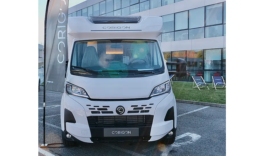 Das Bild zeigt ein Corigon-Wohnmobil stehend von vorne fotografiert. Neben dem teilintegrierten Reisemobil steht ein Werbewimpel mit Corigon-Aufschrift.