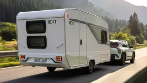 Der Next 381 im Zugbetrieb mit einem Kia EV3 auf einer Landstraße mit Bergen im Hintergrund fahrend von schräg hinten rechts. - Foto: Fendt-Caravan Der Next 381 im Zugbetrieb mit einem Kia EV3 auf einer Landstraße mit Bergen im Hintergrund fahrend von schräg hinten rechts. - Foto: Fendt-Caravan