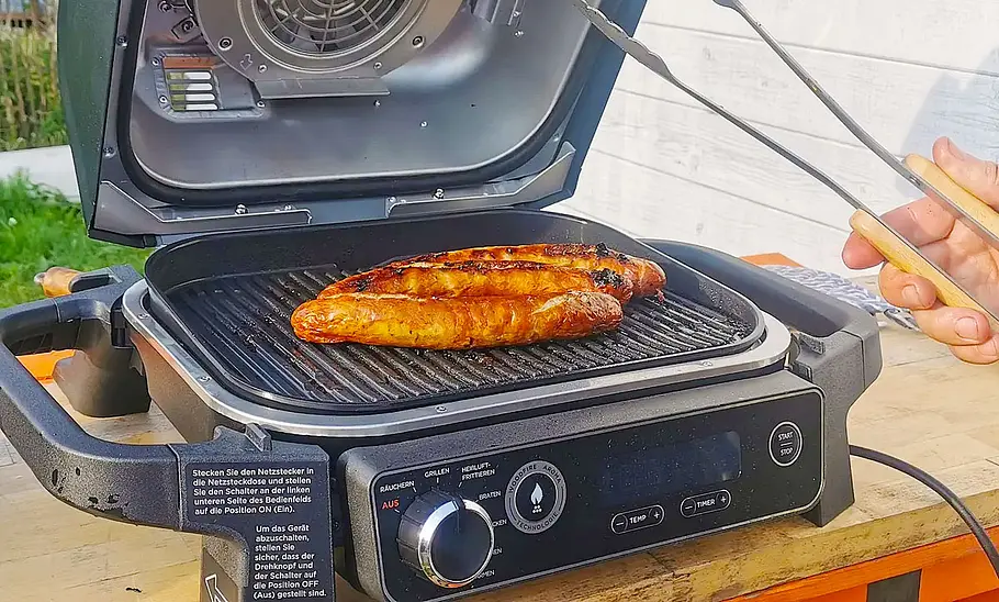 Ninja Woodfire Elektrogrill Der Ninja Woodfire Elektrogrill mit geöffnetem Deckel fotografiert. Es liegen fertig gegrillte Würstchen auf dem Rost.