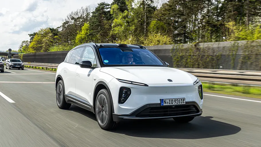 Ein weißer Nio EL6 mit schwarzem Dach fährt auf einer Autobahn. Dahinter sind weitere Autos, eine Lärmschutzwand und Bäume zu sehen.