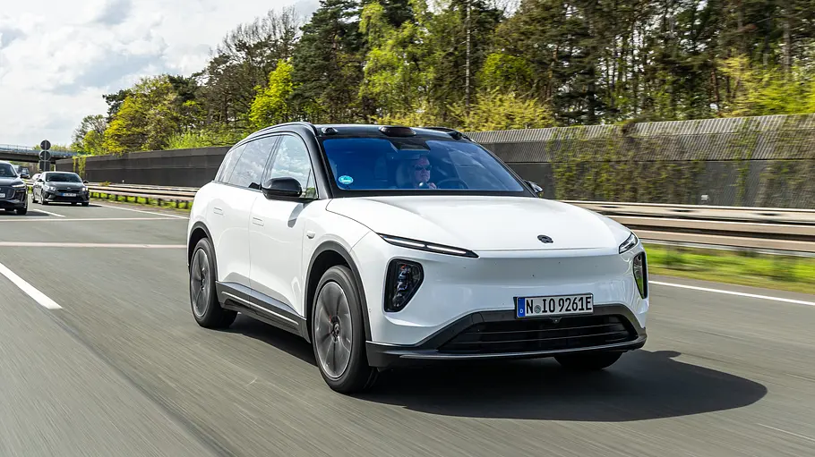Ein weißes Nio EL6 Long Range mit schwarzem Dach fährt auf einer Autobahn. Dahinter sind weitere Autos, eine Lärmschutzwand und Bäume zu sehen.