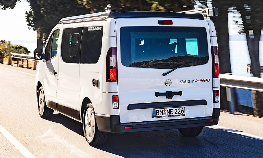 Die Außenaufnahme zeigt den fahrenden Nissan Primastar Seaside by Dethleffs (2022) von schräg hinten.