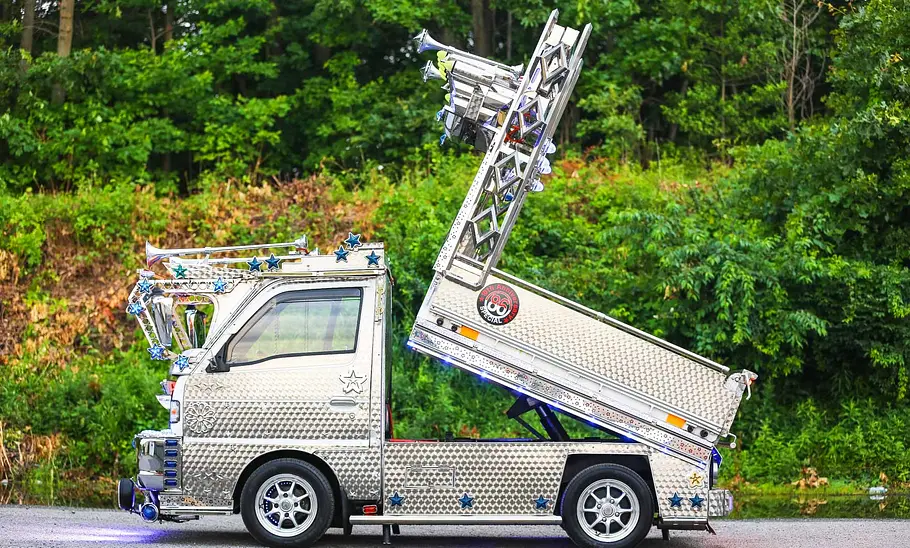Der Suzuki Carry Dekotora steht vor einem Wald und die Ladefläche ist angehoben, seitlich fotografiert.