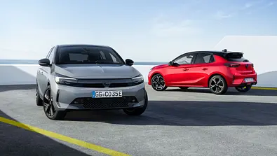 Zwei statische Opel Corsa vor einer weißen Wand. Links in Grau ist der Opel Corsa Electric von schräg vorne rechts, rechts der Opel Corsa Hybrid in Rot von schräg hinten links. - Foto: Opel