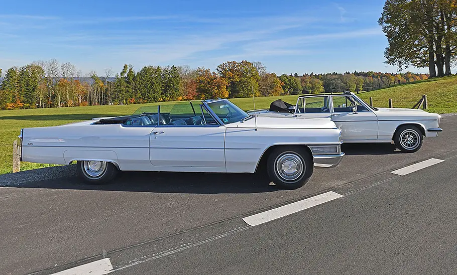 Der Cadillac De Ville Convertible und das Opel Diplomat B Cabriolet stehen vor einer hügeligen Wiesenlandschaft, fotografiert von der Seite.
