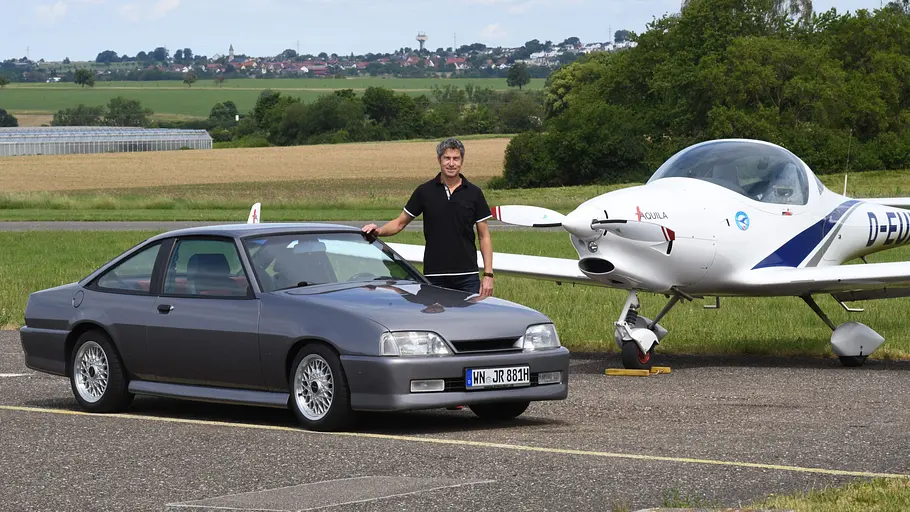Günther Irmscher stehend am Opel Manta C mit Sportflugzeug im Hintergrund.
