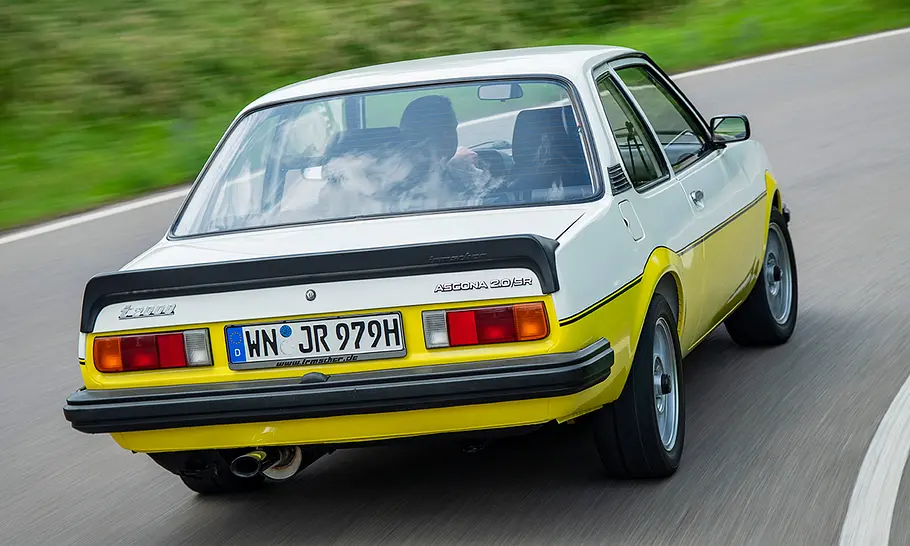 Der Opel Ascona B i 2000 fährt auf einer Straße, fotografiert von hinten schräg.