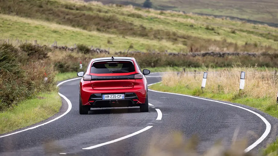Peugeot E-208 GTi Fahrbericht Ein roter Peugeot e-208 GTi ist von hinten auf einer kurvigen Landstraße in einer grünen, grasbewachsenen Hügellandschaft zu sehen.