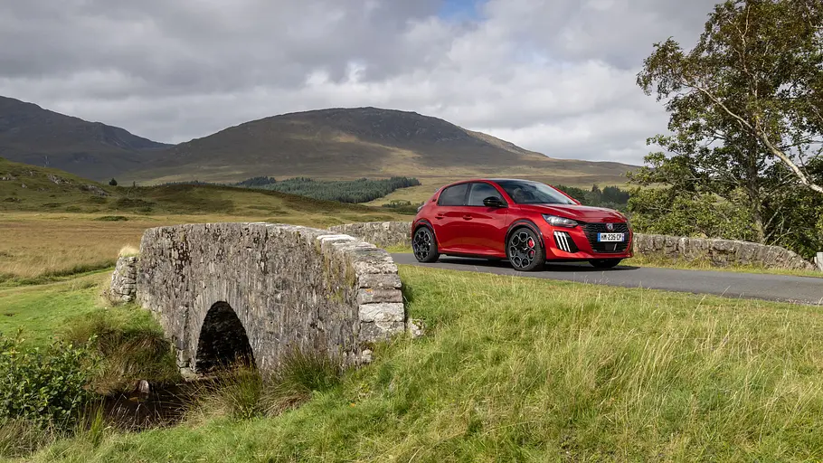 Peugeot E-208 GTi Fahrbericht Ein roter Peugeot E-208 GTi fährt über eine steinerne Bogenbrücke in einer grünen Berglandschaft unter bewölktem Himmel.