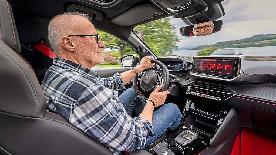 Peugeot E-208 GTi Fahrbericht Ein Mann mit Brille und kariertem Hemd sitzt am Steuer des Peugeot E-208 GTi. Durch die Frontscheibe sind ein See und Hügel sichtbar.