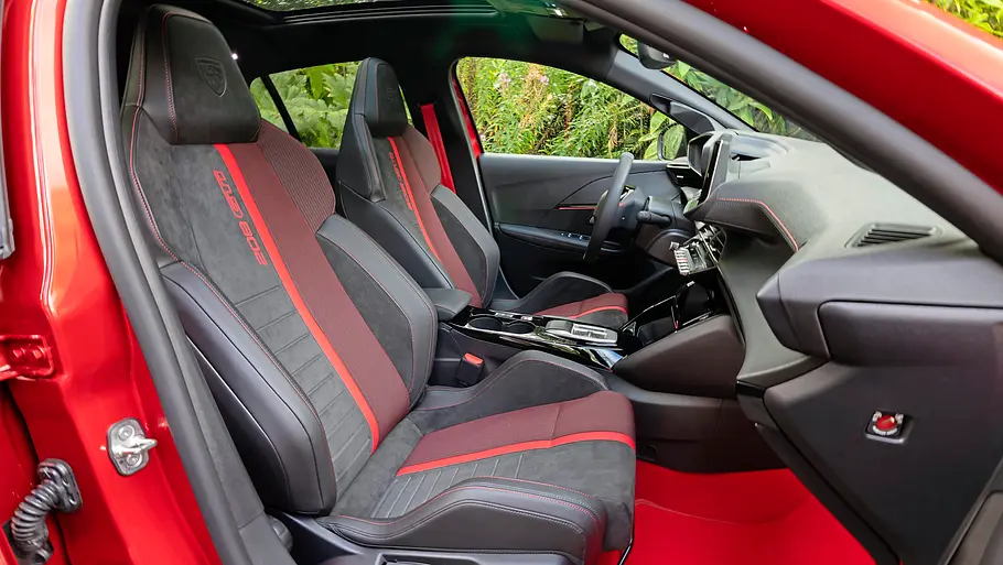 Peugeot E-208 GTi Fahrbericht Der Innenraum eines roten Peugeot E-208 GTi zeigt schwarz-rote Sportsitze, rote Fußmatten, ein dunkles Armaturenbrett und ein Glasdach.
