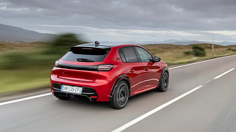 Peugeot E-208 GTi Fahrbericht Ein roter Peugeot E-208 GTi fährt auf einer asphaltierten Straße durch eine karge Berglandschaft unter bewölktem Himmel.