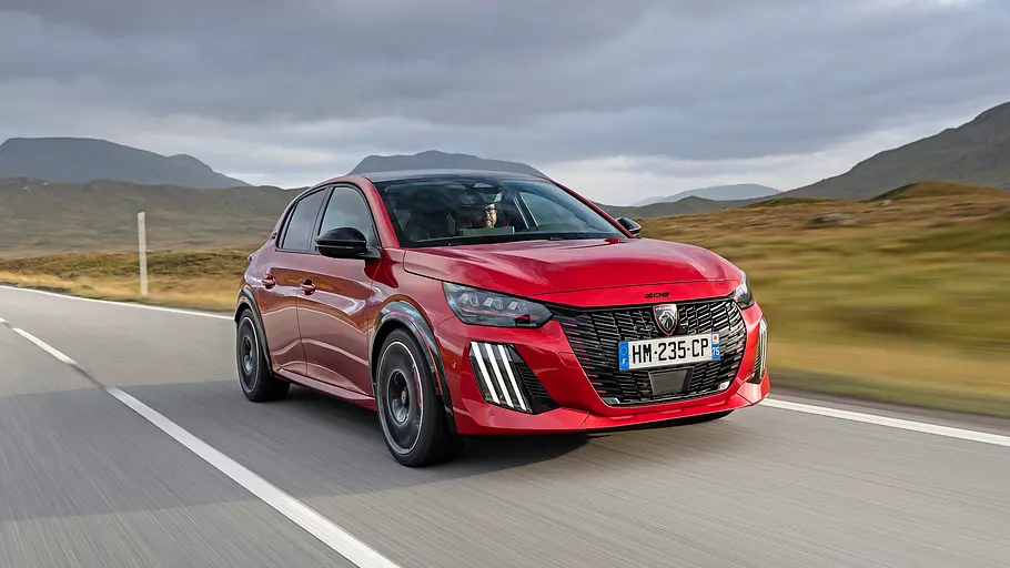 Peugeot E-208 GTi Fahrbericht Ein roter Peugeot E-208 GTi fährt auf einer asphaltierten Straße durch eine karge Berglandschaft unter bewölktem Himmel.
