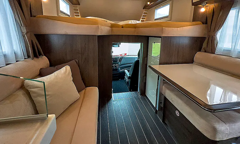 Phoenix Midi Alkoven 6700 HBL (2025) Das Bild zeigt den Wohnraum des Phoenix Midi Alkoven 6700 HBL (2025). Zu sehen ist die Sitzgruppe. Der Zugang zum Fahrerhaus ist geöffnet.