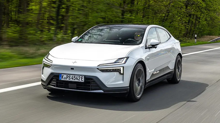 Ein weißer Polestar 4 Long Range mit schwarzem Dach fährt auf einer Straße an einem grünen Wald vorbei, schräg von vorn gesehen.